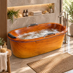 Oval wood-look freestanding tub with non-slip grab bars and black headrest, set in warm stone bathroom with floor faucet, green plants and folded towels
