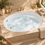 Oval drop-in bathtub in sunlit bathroom with beige marble surround, accessorized with toiletries, towels and green plants