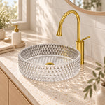 Round diamond embossed clear glass vessel sink with gold faucet installed on speckled vanity countertop in bright modern bathroom