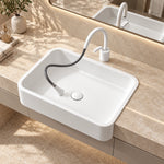 White ceramic semi-recessed bathroom sink with rounded rectangular basin and flexible pull-out faucet on a beige stone countertop.